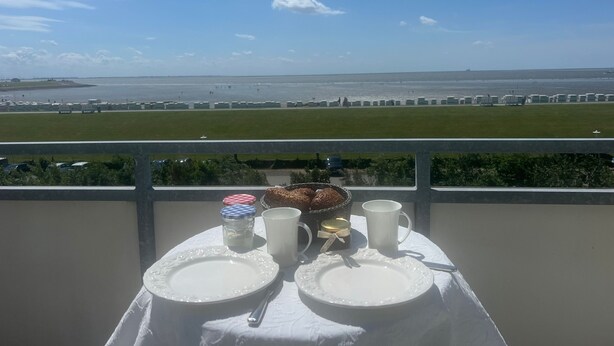 Mesa posta com pratos, caneca e cesta, numa varanda com vista para a praia e o aeroporto.