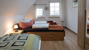 Une chambre avec un lit en mezzanine, une table de chevet avec une lampe, deux fenêtres avec des rideaux et un radiateur.