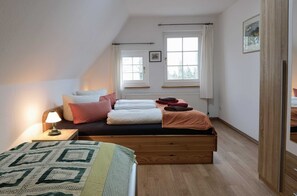 Un dormitorio con cama doble superior, una mesita de noche con lámpara, dos ventanas con cortinas y un radiador.