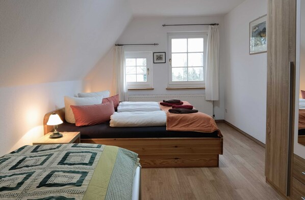 Een slaapkamer met een hoogslaper, een nachtkastje met lamp, twee ramen met gordijnen en een radiator.