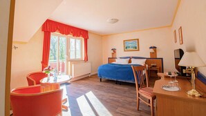 Ein Hotelzimmer mit einem Bett, einem Schreibtisch mit Lampe, einem Sessel, einem Tisch mit Blumenvase und einem Fenster mit roten Vorhängen.