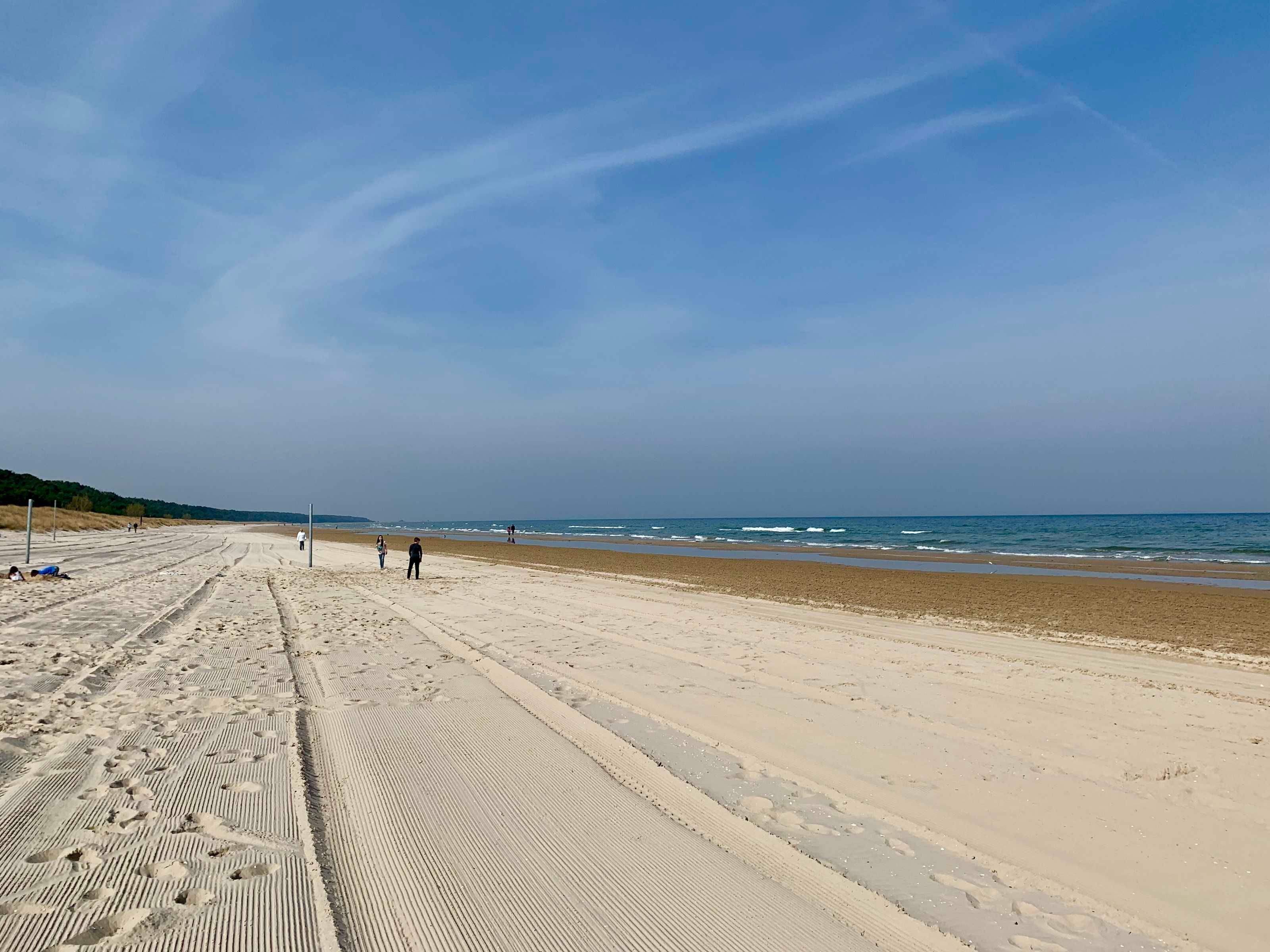 A wide sandy beach with footprints and tire tracks, a clear sky, and a calm sea.