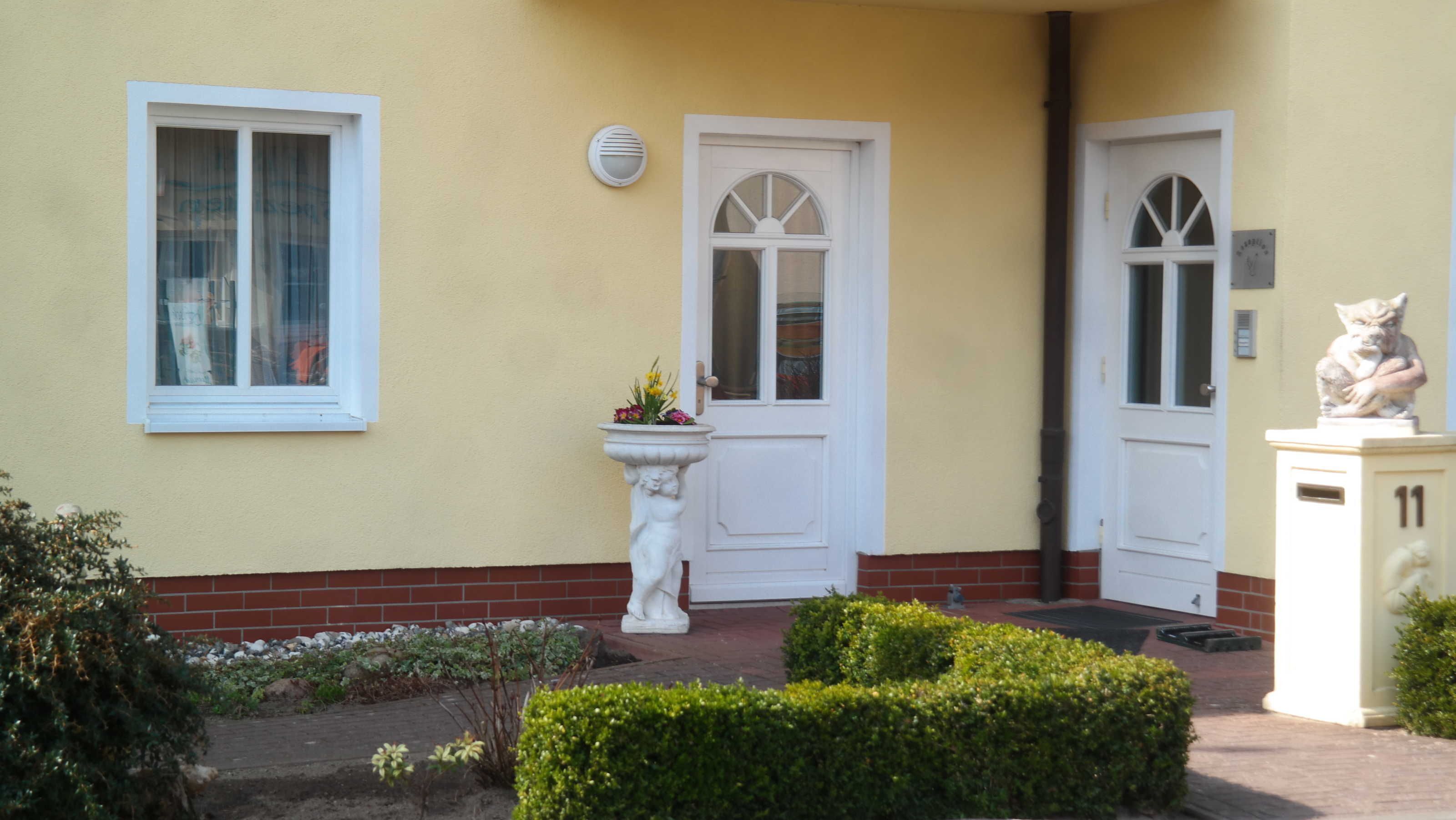 A building with two white doors, a window with a white frame, a white wall-mounted light, and a small garden with a statue and a planter.