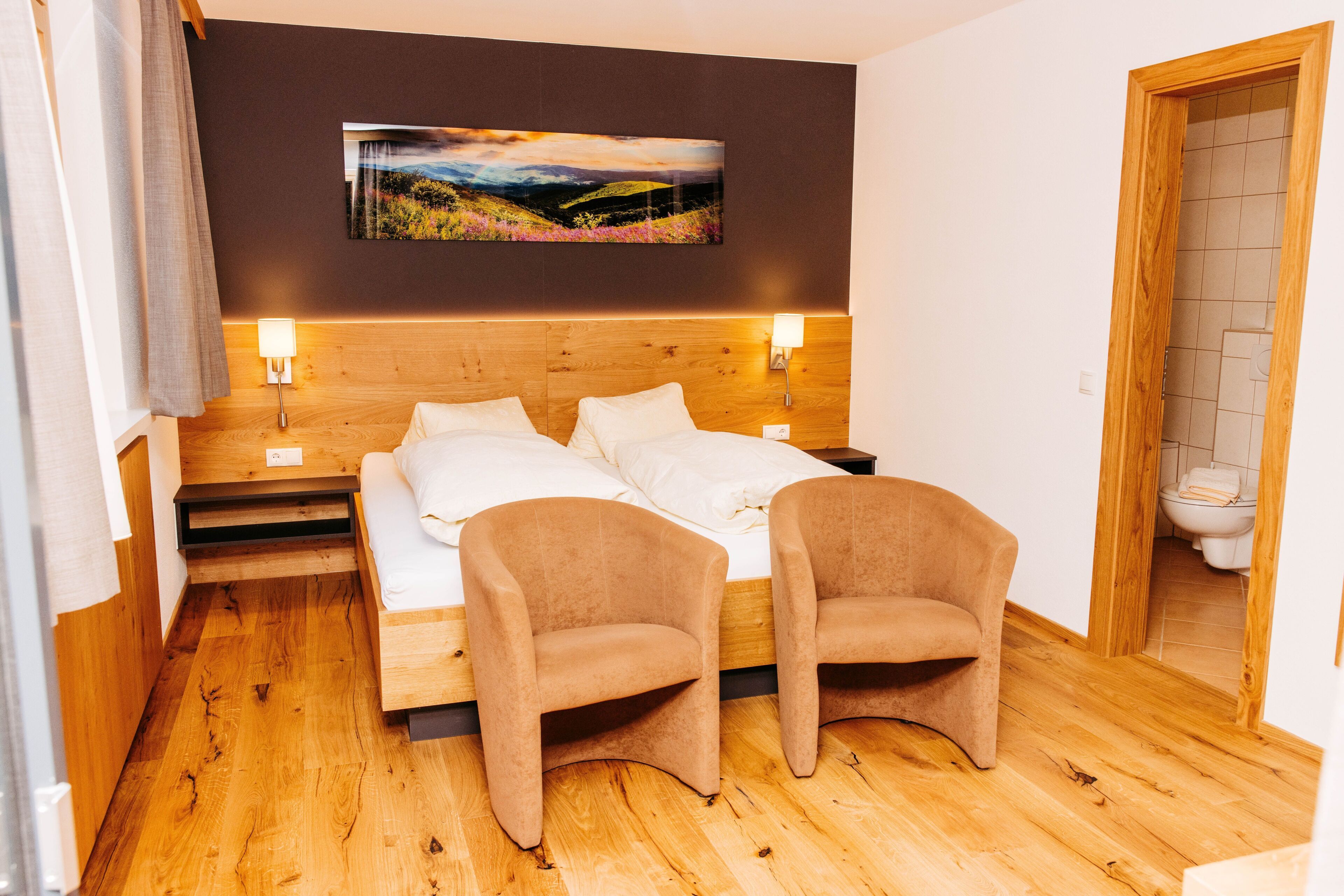 Ein Hotelzimmer mit einem Holzkopfteil, einem Bett mit weißen Leinen, zwei Sesseln, einem Bild an der Wand und einem Holzboden.