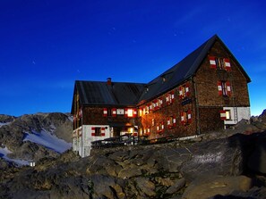 Un grand chalet de montagne rustique, doté de nombreuses fenêtres et d’un toit à forte pente, entouré d’un paysage rocheux et enneigé.