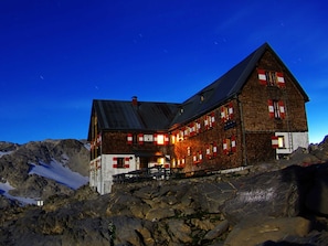 A large, rustic mountain lodge with multiple windows and a steep roof, surrounded by rocky terrain and snow.