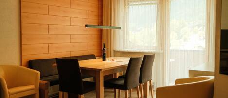 A dining area with a wooden table and chairs, a sofa, and a window with sheer curtains.