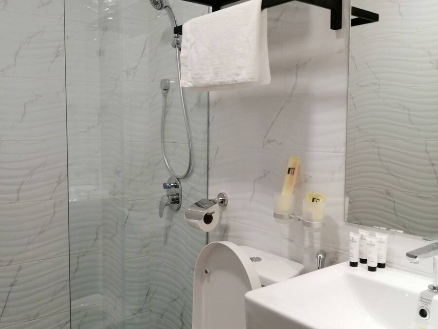 Moderni kylpyhuone, jossa on lasiseinällä varustettu suihku, valkoinen wc-istuin ja pesuallas, jonka päällä on hygieniatuotteita.