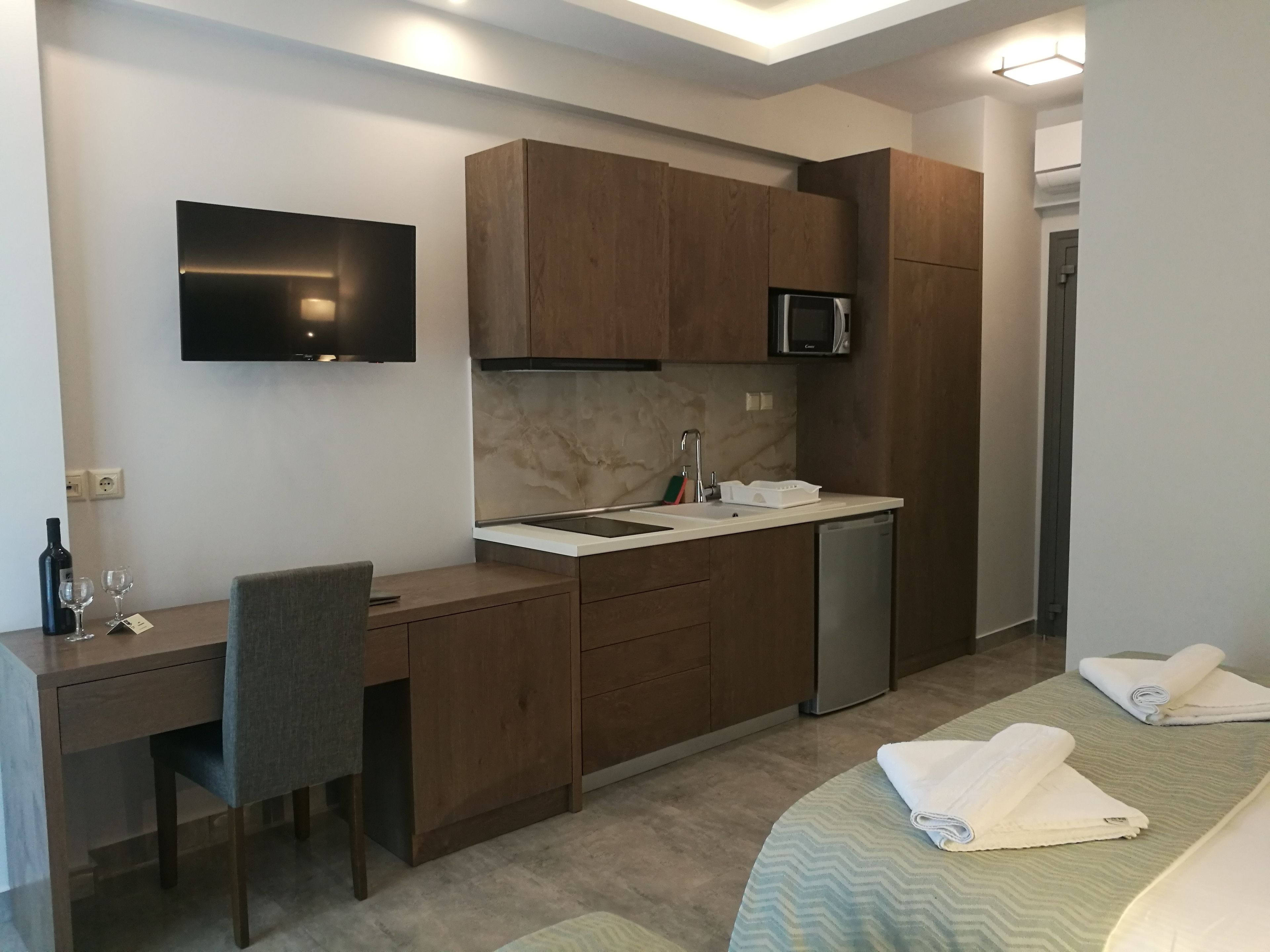 A compact hotel room with a kitchenette, a flat-screen TV, a bed with towels, and a desk with a chair.