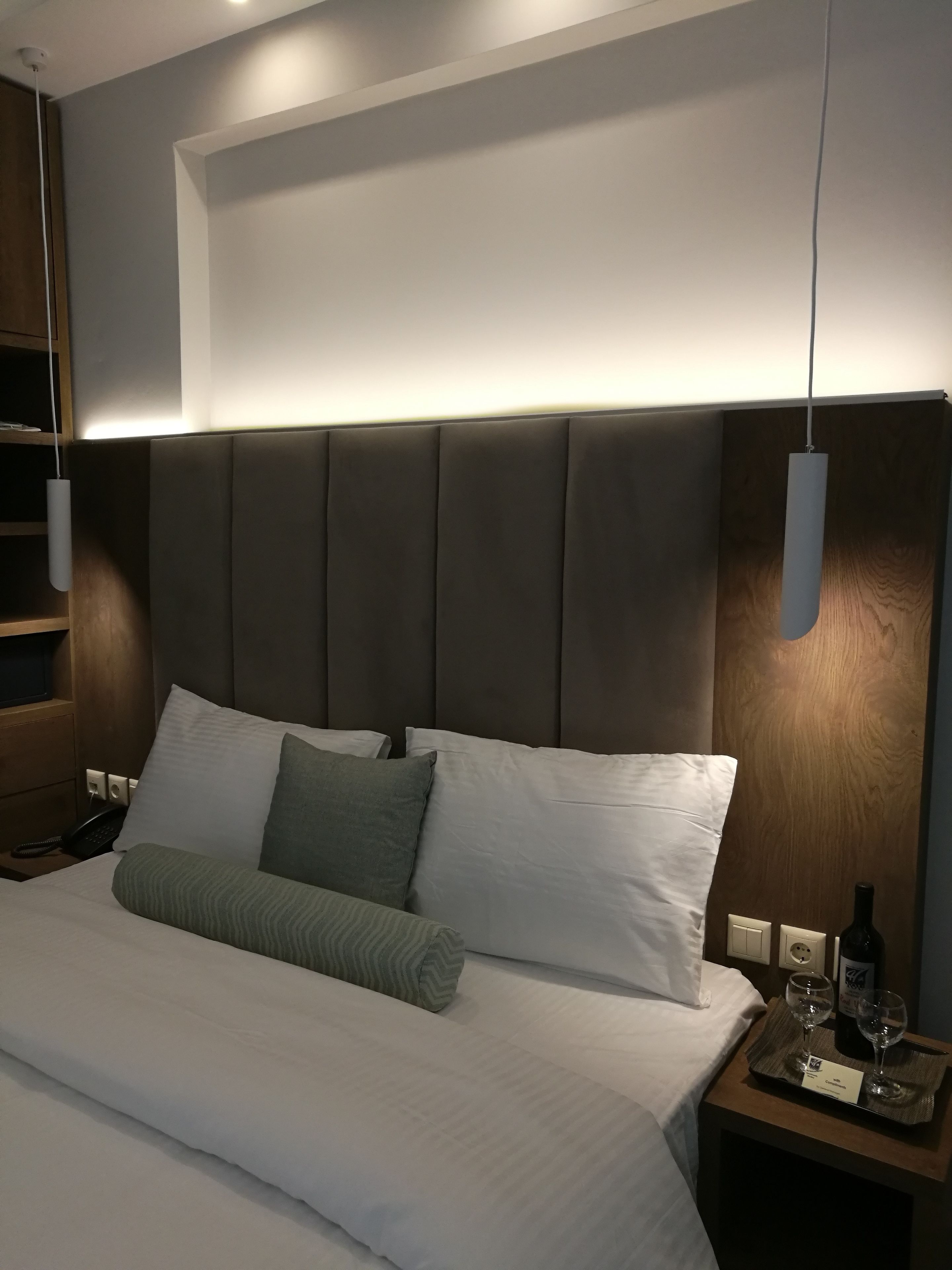 A neatly made bed with white and green pillows, a bedside table with a wine bottle and glasses, and a modern headboard.