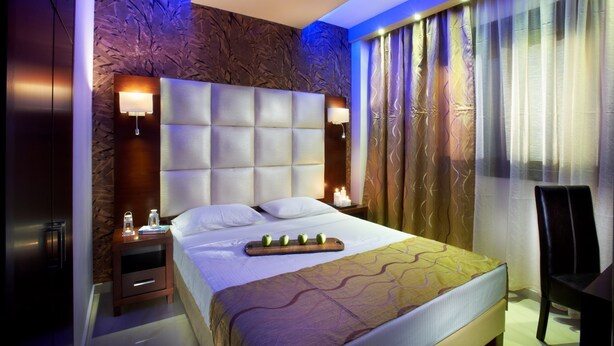 A modern hotel room with a large bed, a headboard with a light panel, a nightstand with a lamp, and a view of the outside through sheer curtains. - 4-You Family 2 (Polygyros)