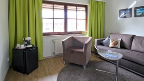 Ein modernes Wohnzimmer mit einem grauen Sofa, einem kleinen runden Tisch und einem braunen Sessel. Unter einem Fenster mit grünen Vorhängen ist eine Heizung sichtbar.