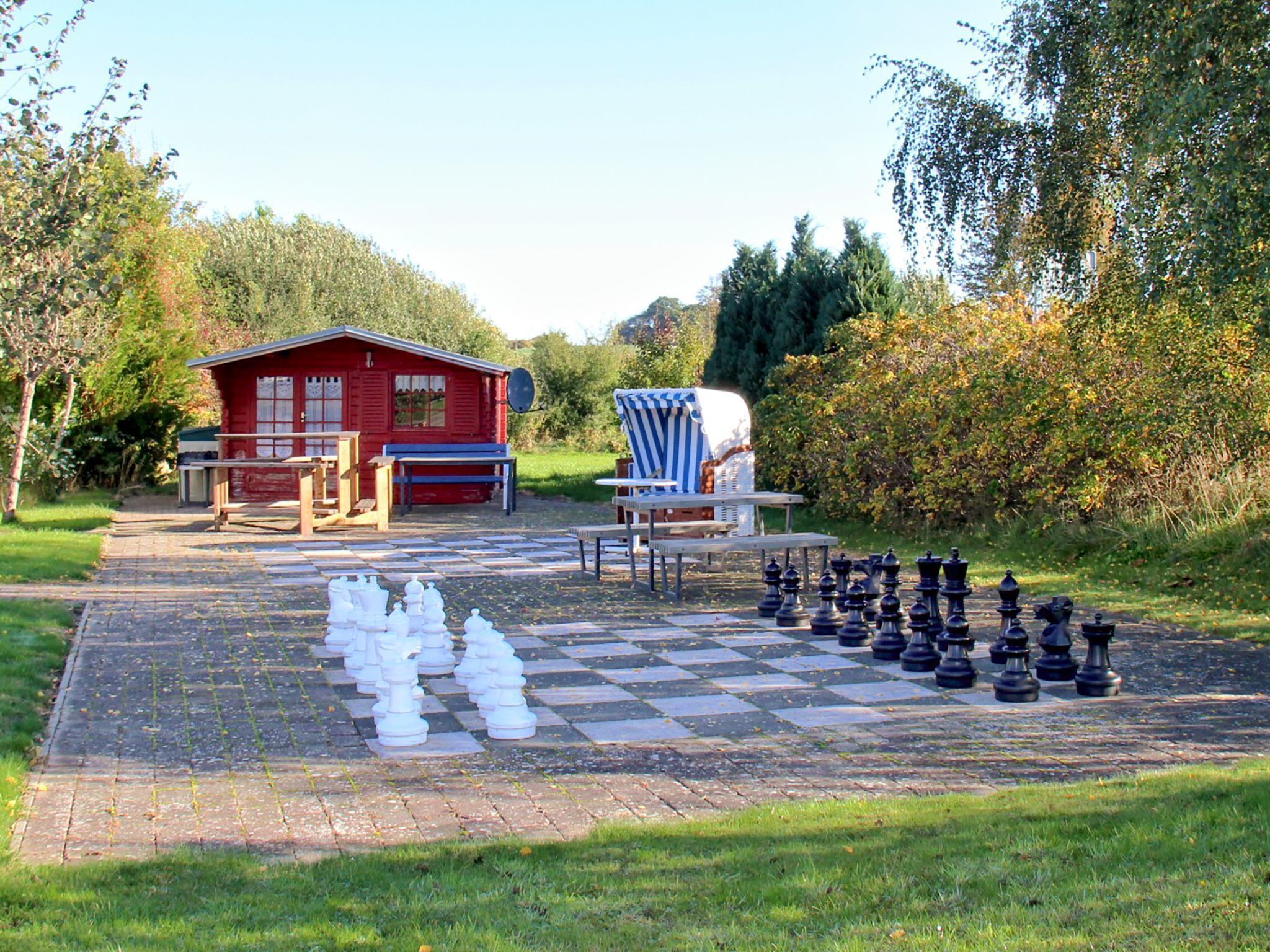 Ein Schachbrett mit kariertem Muster im Freien, aufgestellt in einem Park, mit einem roten Gebäude und einem blau gestreiften Stuhl im Hintergrund.