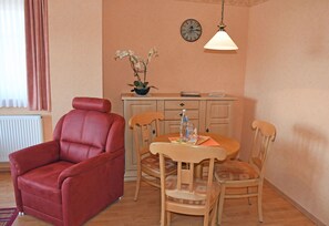 Ein gemütliches Wohnzimmer mit einem roten Sessel, einem Holzespeisetisch, Stühlen, einer kleinen Kommode und einer Uhr an der Wand.