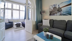 A modern living room with a grey sofa, a blue armchair, and a white coffee table. There is a painting of a boat on the wall and a window with curtains. - 05 Ferienappartement Jasmund (H) - Apartment house Haus Arkona (Sellin)
