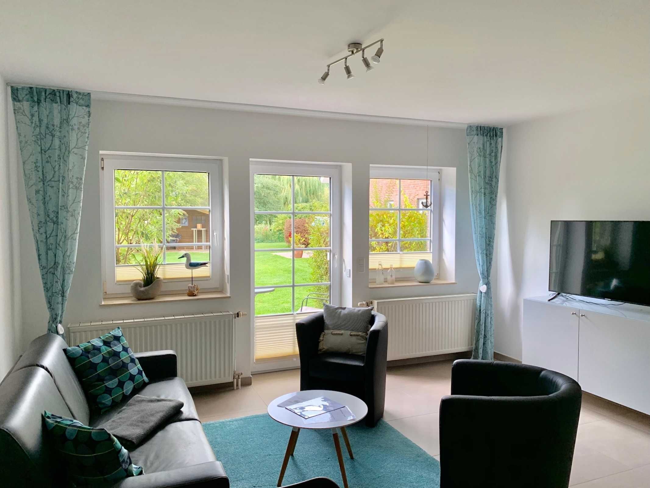 A modern living room with a sofa, two armchairs, a small table, and a TV. There are three large windows providing views of a garden.