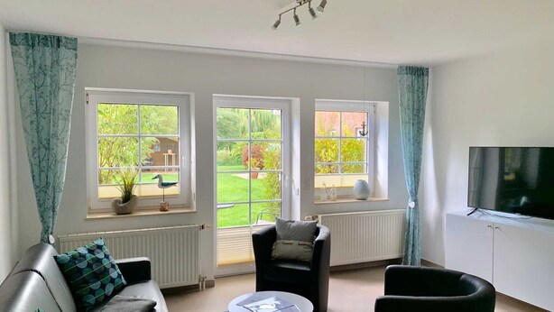 Ein modernes Wohnzimmer mit einer Couch, zwei Sesseln, einem kleinen Tisch und einem Fernseher. Drei große Fenster bieten Blick auf einen Garten.