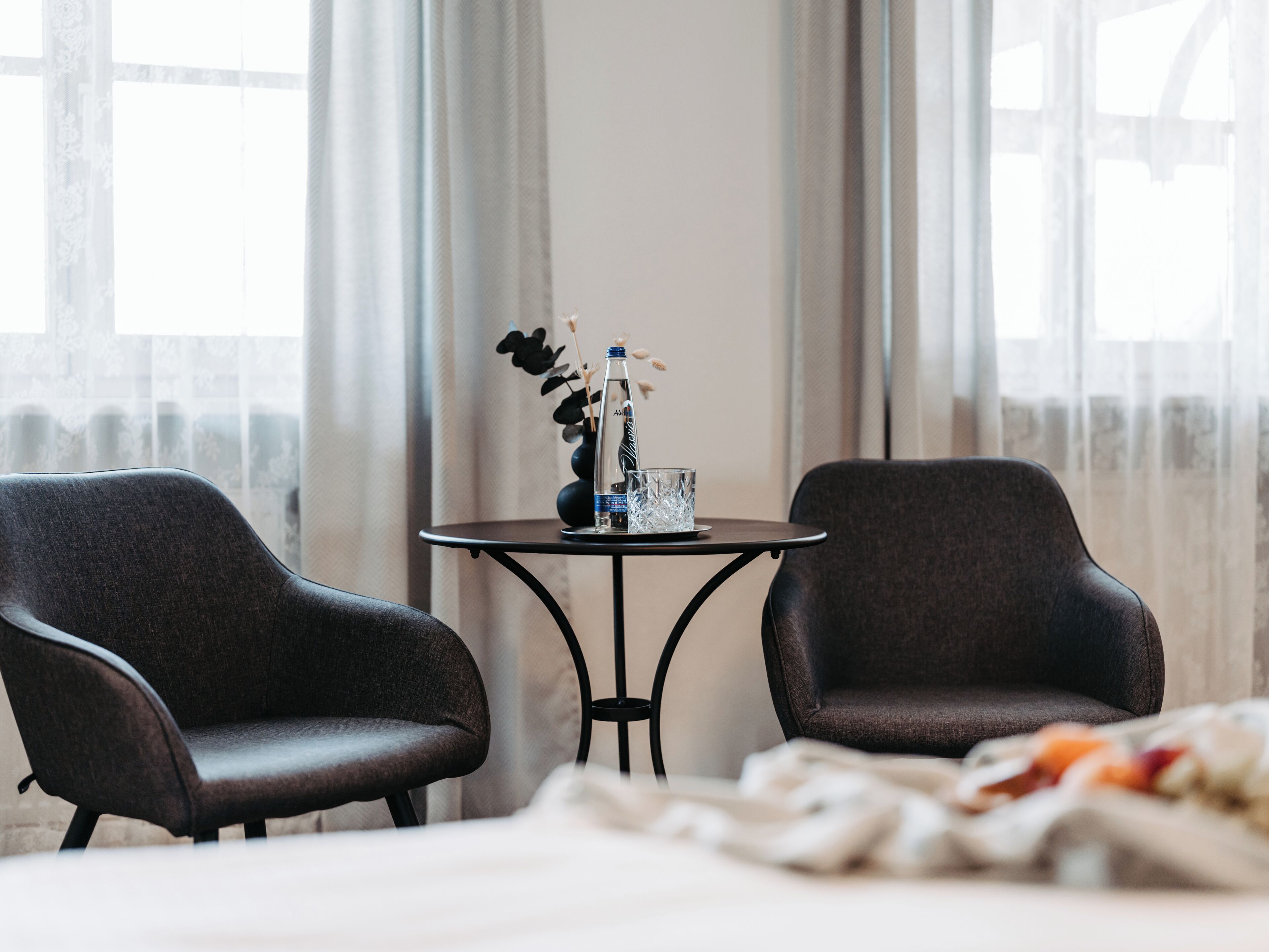 A hotel room with two dark armchairs, a small round table with a vase, and a bed with a white bedspread.