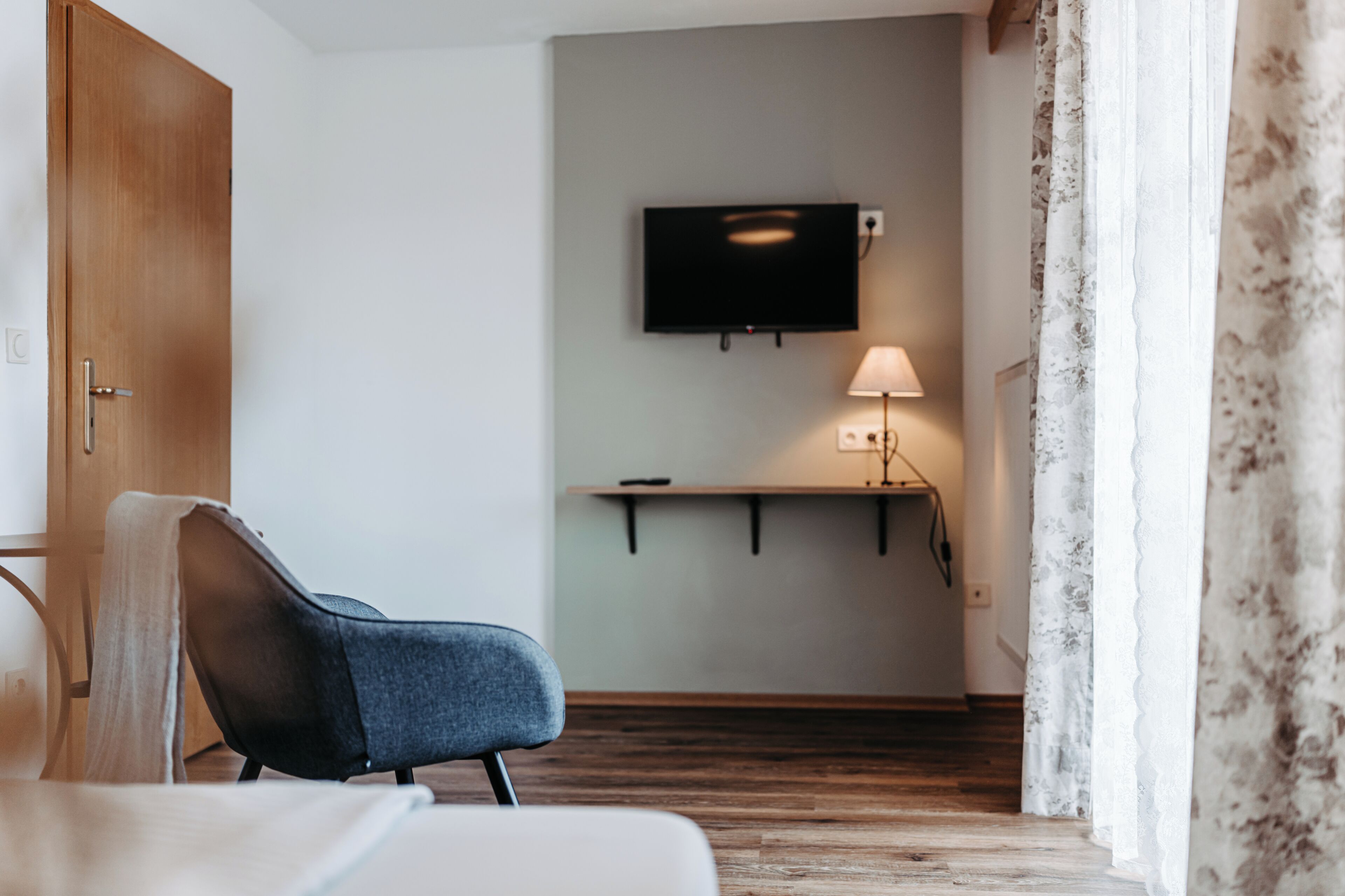 Une chambre d’hôtel avec une porte en bois, un téléviseur à écran plat fixé au mur, une table de chevet avec une lampe et une chaise avec une couverture.