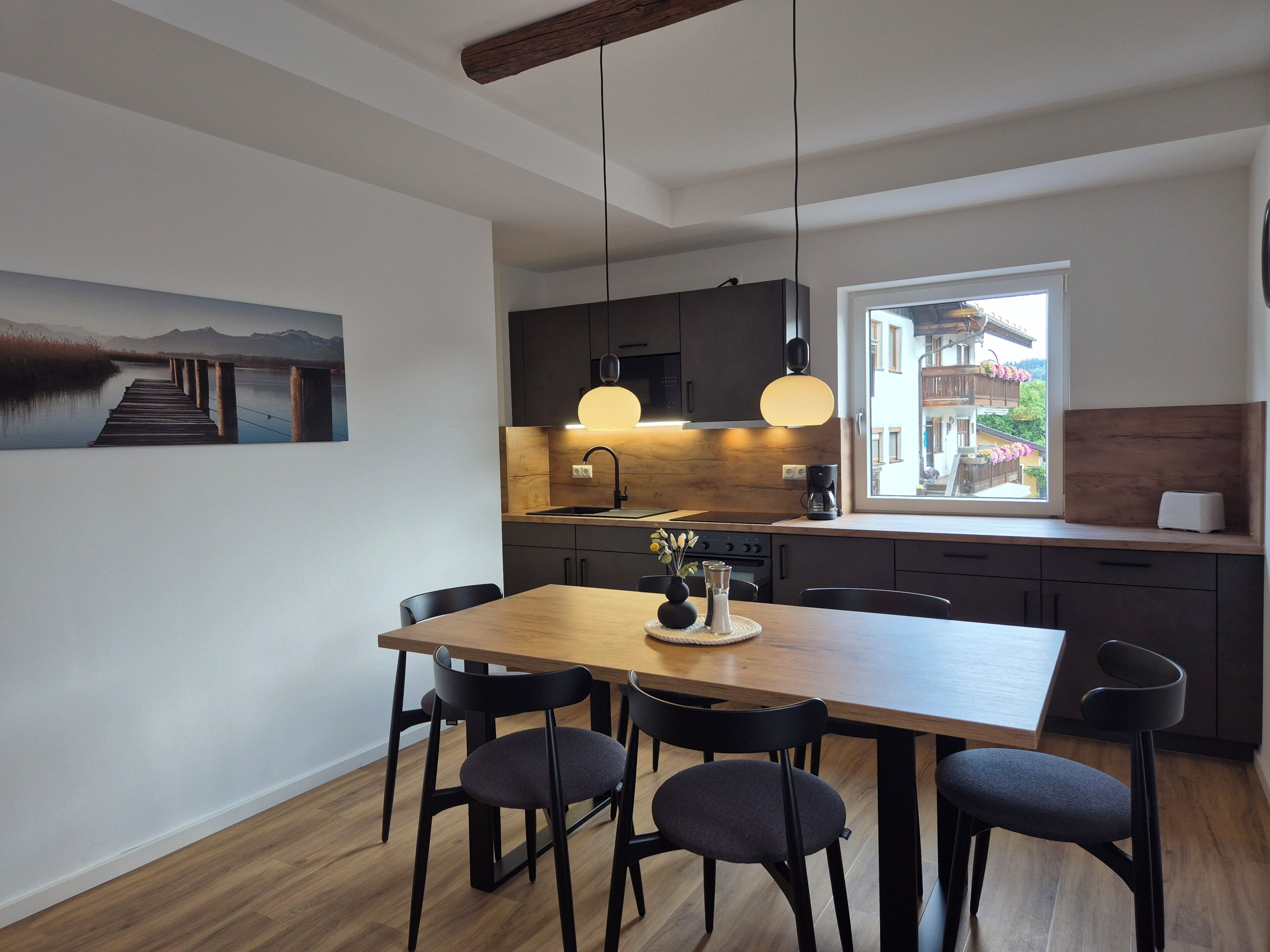 Une cuisine moderne avec une table à manger en bois, des meubles de rangement sombres et une fenêtre donnant sur un immeuble.
