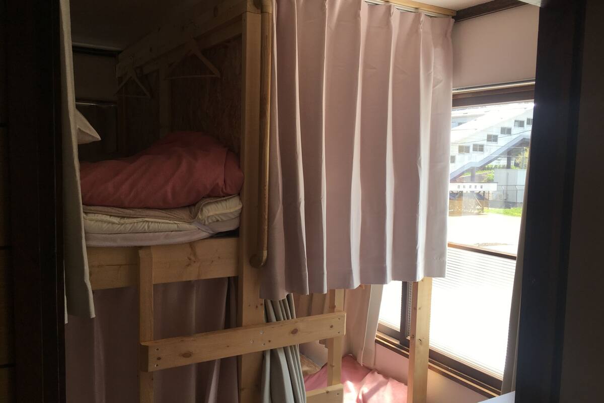 Una cama litera con colchón rosa, escalera de madera y cortina entreabierta.