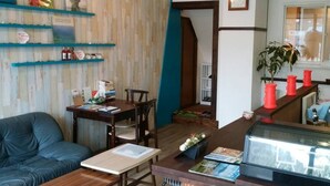 Een gezellige zithoek met een blauwe bank, een houten salontafel en een eettafel met stoelen. Er zijn boekenkasten met boeken en decoratieve items, een klok aan de muur en een raam met uitzicht.