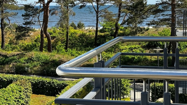 Une rambarde métallique surplombe un espace paysager agrémenté d’arbres et offrant une vue sur la mer.