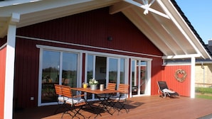 A red building with a covered patio area featuring a wooden table and chairs, a glass door leading inside, and a small outdoor seating area with a lounge chair.
