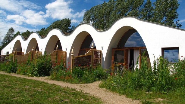 A row of white, wavy-roofed cabins with wooden doors and windows, surrounded by lush greenery and a clear blue sky.