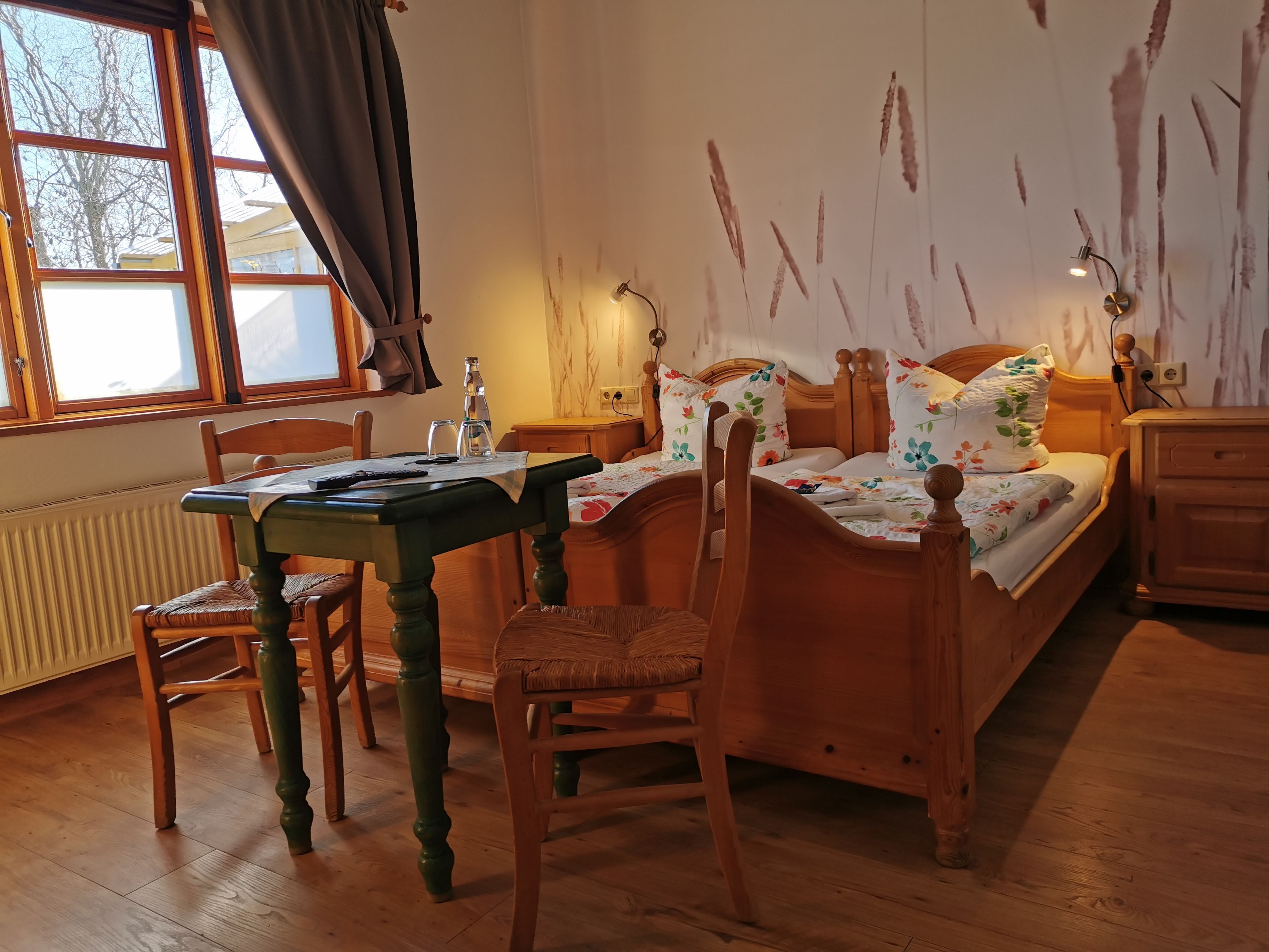 A room with a wooden bed, a wooden table, a chair, a window with curtains, and a radiator.