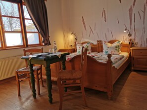 A room with a wooden bed, a wooden table, a chair, a window with curtains, and a radiator. - 2 - "Up'n Hoff", Germany (Middelhagen)