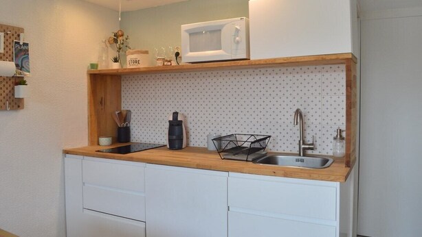 A compact kitchen with white cabinets, a wooden countertop, a microwave, a sink, and a small shelf with kitchen items.