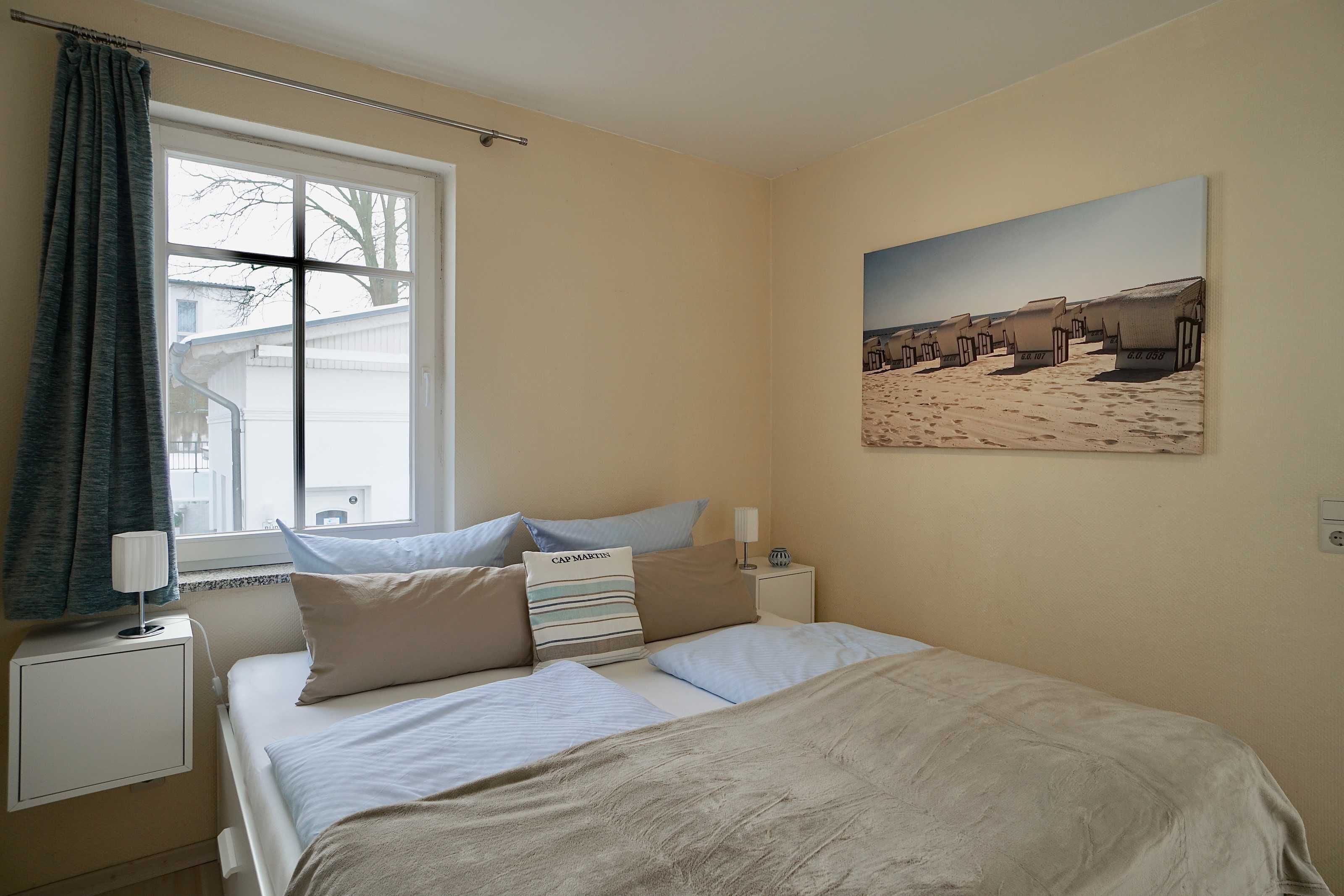 Een slaapkamer met een bed, nachtkastje, lamp, raam met gordijnen en een schilderij van een strand.