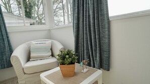A room with a white sofa, a small wooden table with a plant, and a view of trees through a window. - 08 Ferienappartement Mönchgut (H) - Apartment house Haus Arkona (Sellin)