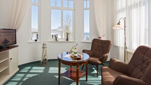 Una habitación con un ventanal grande, un televisor, una mesita con un objeto decorativo, un sillón marrón y una lámpara de pie.