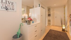 Un couloir avec une commode blanche, un miroir, une pancarte sur laquelle on peut lire « VIVEZ VOS RÊVES », et un parapluie vert dans un panier.