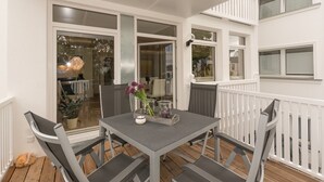 Une terrasse extérieure avec une table et des chaises, une balustrade blanche et une vue sur l’intérieur de la maison.