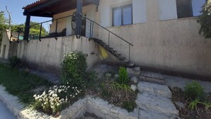Uma casa com uma escadaria de pedra que leva a um deck de madeira, uma parede de concreto e um pequeno jardim com flores brancas e plantas verdes.