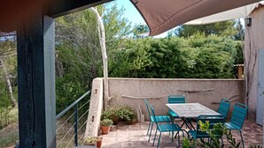 Pátio externo com mesa e cadeiras, rodeado por plantas em vasos e vista para árvores.
