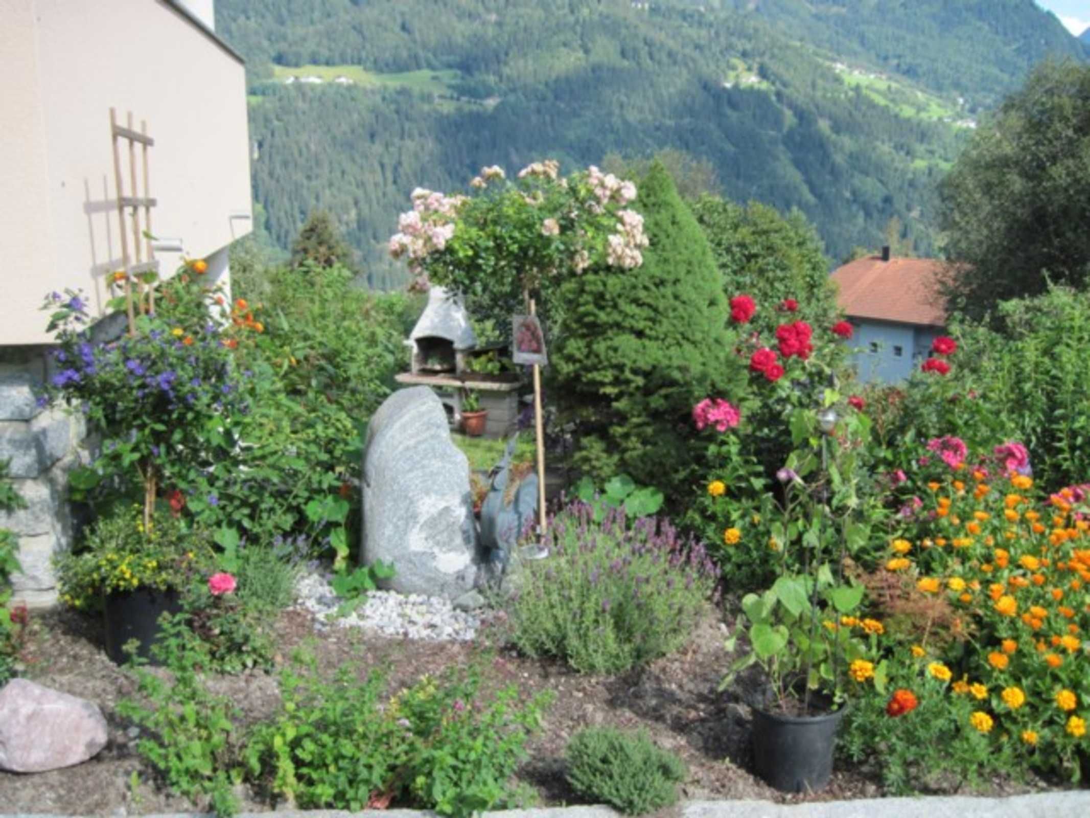 Un jardin avec différentes plantes et fleurs, une structure en pierre et un bâtiment avec un toit rouge.