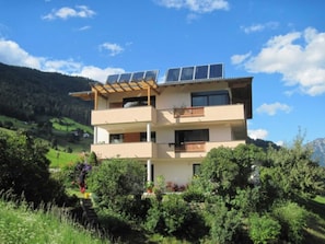 Un immeuble à plusieurs étages, doté de panneaux solaires sur le toit, entouré de verdure et de collines.