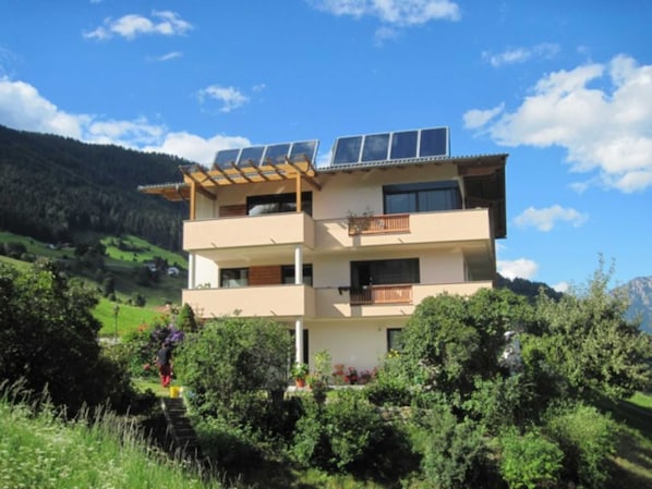 A multi-story building with solar panels on the roof, surrounded by greenery and hills. - Bauernhof Huber - Double room 1 (Wenns)