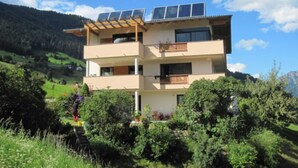 Un immeuble à plusieurs étages, doté de panneaux solaires sur le toit, entouré de verdure et de collines.