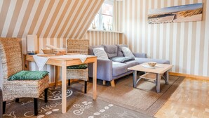 Ein gemütliches Zimmer mit Streifenmuster an den Wänden, einem Holztisch, einer Couch und Blick auf den Strand durch ein Fenster.