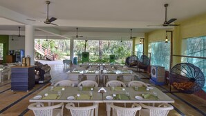 Un vaste espace extérieur aménagé pour les repas, avec plusieurs tables et chaises, des ventilateurs de plafond et une vue sur la verdure environnante.