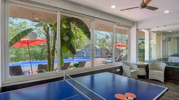 A ping pong table with two paddles, a ceiling fan, and a view of an outdoor area with red umbrellas and chairs.