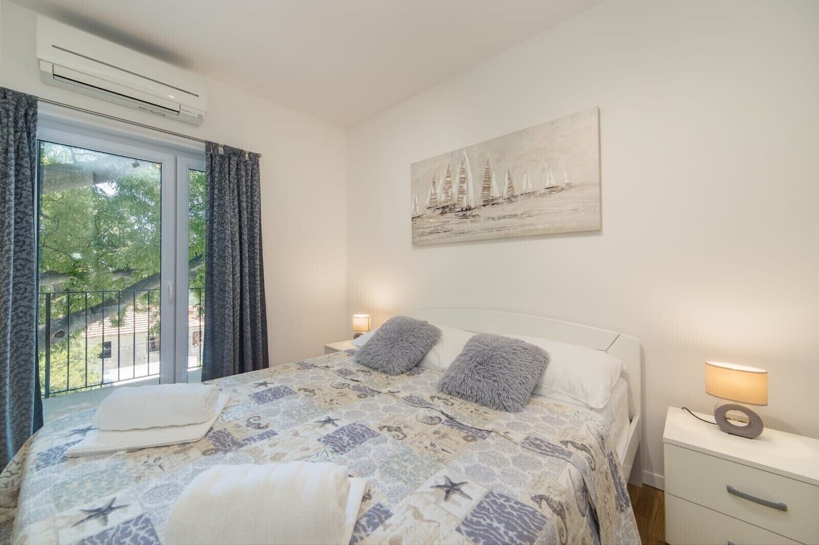 A bedroom with a bed, a window with curtains, a painting of sailboats, and a nightstand with a lamp.