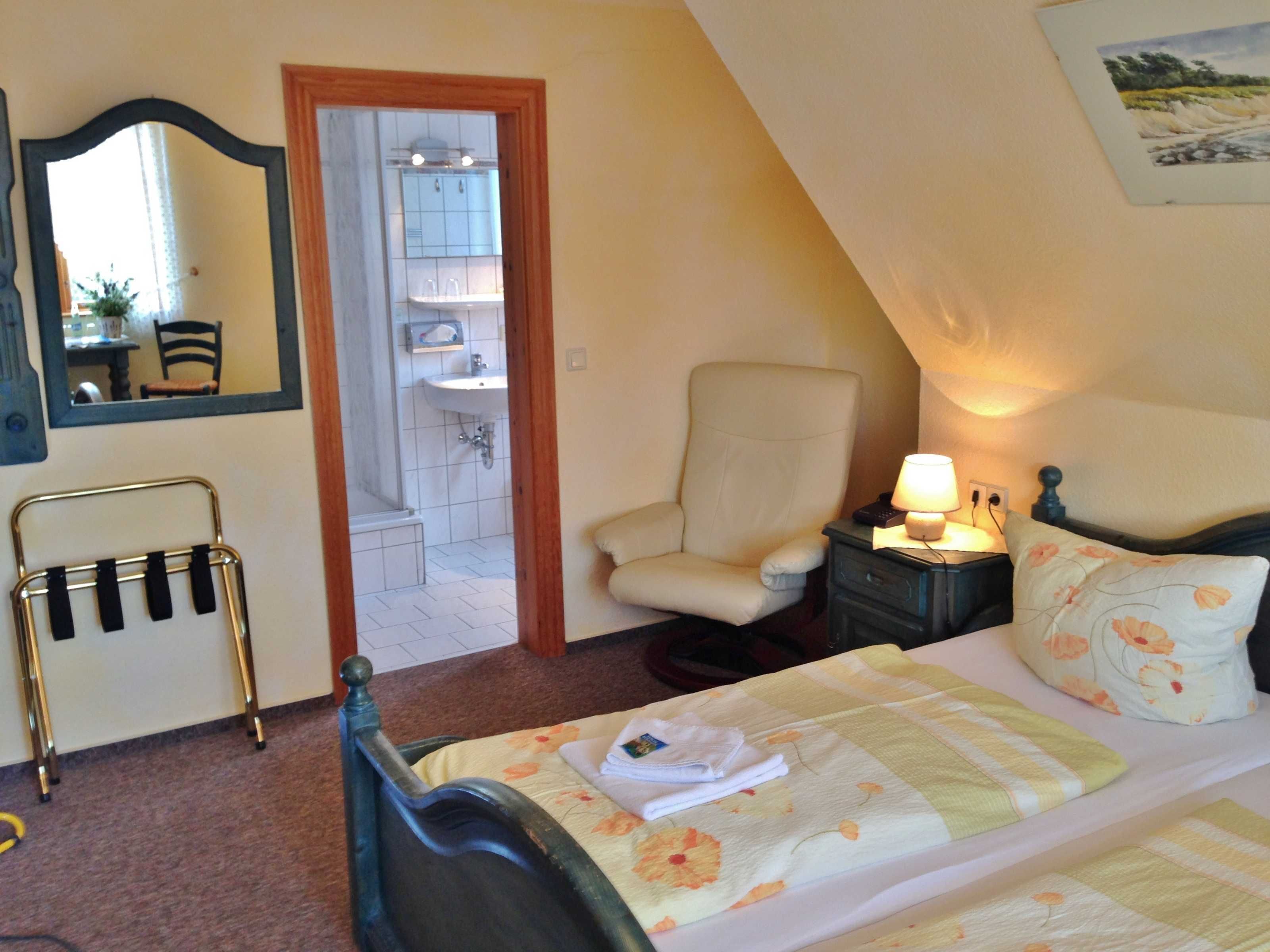 A bedroom with a bed, a chair, a nightstand with a lamp, a mirror, and a view of the bathroom.
