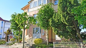 A two-story building with a beige facade, a visible entrance with a door and steps, and a small garden area with potted plants and a bench.