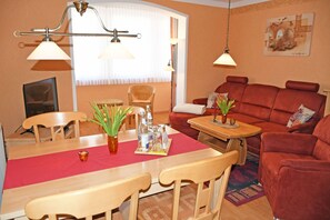 Uma área de refeições com uma mesa de madeira, cadeiras e um jogo americano vermelho. Há uma cadeira de vime e um sofá vermelho ao fundo.